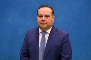 A man wearing a blue suit, light blue shirt, and blue textured tie stands in front of a plain blue background, reflecting the professionalism often found in SUNY Niagara Board of Trustees: Governing Policies and Structure. He has short dark hair and a neutral expression.