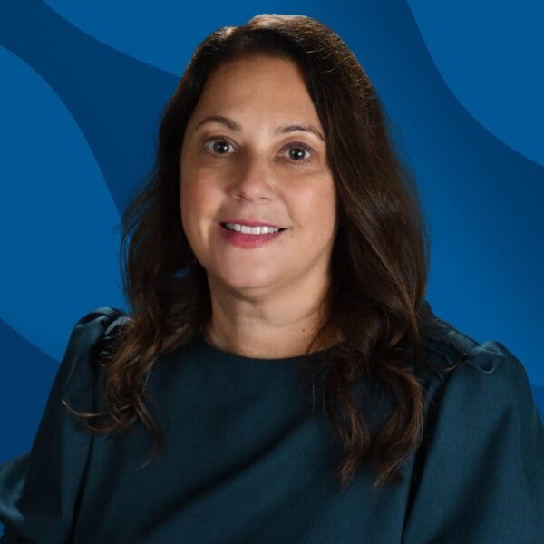A woman with long brown hair wearing a dark teal blouse smiles at the camera. The background features a blue abstract pattern.