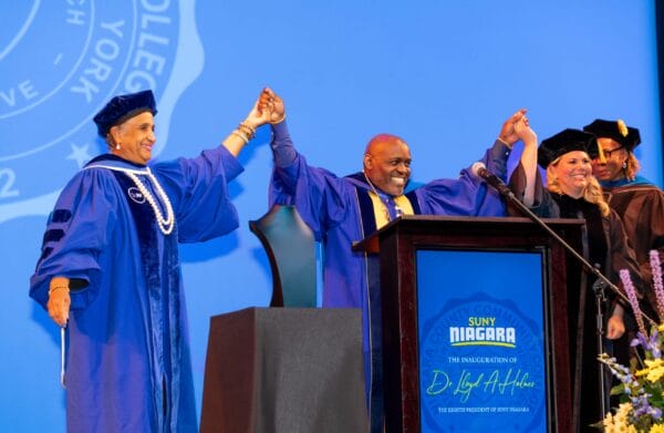 SUNY Niagara's president Dr. Holmes smiling at a podium in blue and yellow graduation robes around flowers holding hands with two people