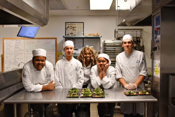 Five people in chef uniforms stand smiling behind a counter with prepared salads in a professional kitchen. A woman in the center wears no chef hat, while the others wear white hats and jackets. Kitchen equipment is visible in the background.