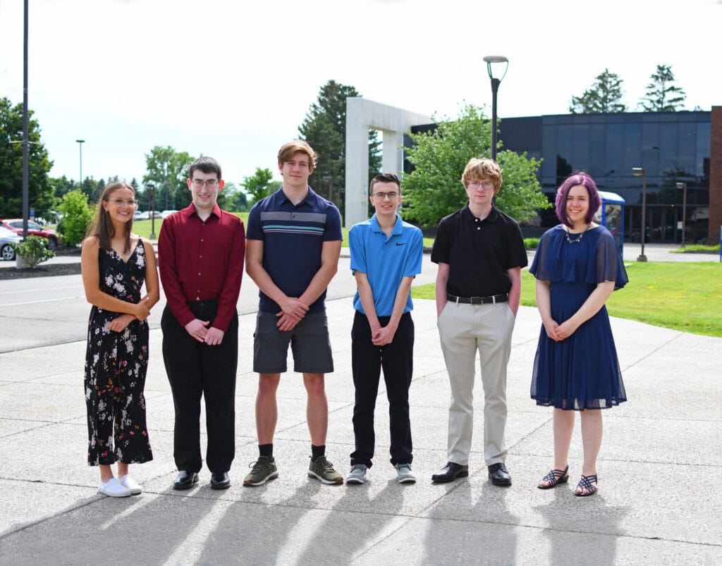 Six young adults stand in a row outside on a sunny day, dressed in semi-formal attire, smiling toward the camera. There are buildings, trees, and a lamp post in the background.