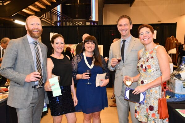 Five people dressed in semi-formal attire stand together at an indoor event, smiling and holding drinks and papers, with tables and a staircase visible in the background.