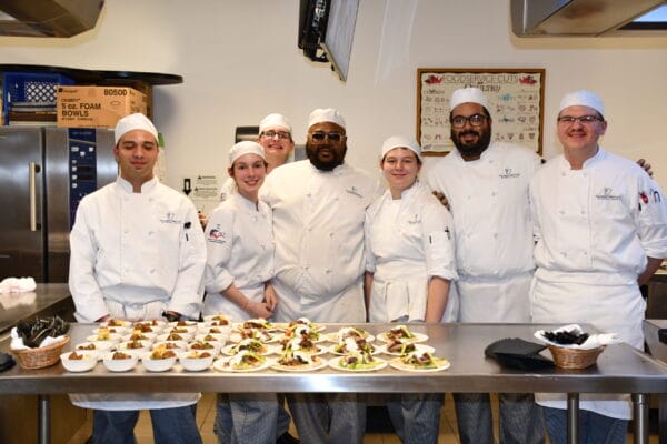 Seven chefs in white uniforms and hats stand together smiling behind a stainless steel counter with several plated dishes and baskets, in a professional kitchen setting.