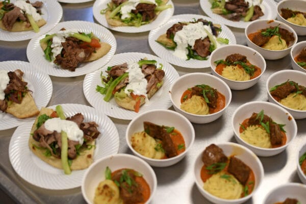 Plates of flatbread topped with sliced meat, vegetables, and white sauce sit beside bowls of mashed potatoes and meat in sauce, arranged on a metal surface, ready to be served.