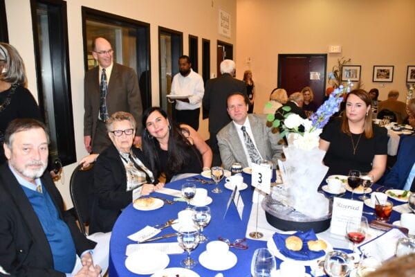 A group of people sits around a round table with blue tablecloths at a formal event, dressed in suits and dresses. There are plates, glasses, and flower centerpieces on the table; other guests and staff are visible in the background.