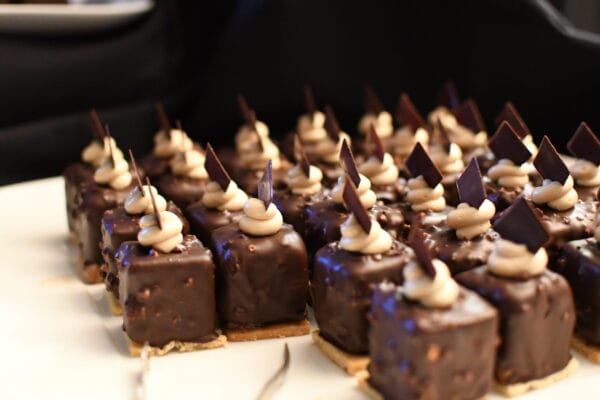 Rows of small, square chocolate desserts topped with a swirl of cream and a dark chocolate shard, arranged neatly on a white tray with a blurred dark background.