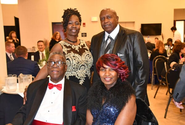 Four adults pose together at a formal event; two men in suits and two women in elegant dresses, with tables and other guests visible in the background.