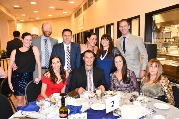 A group of eleven adults dressed in formal attire smile for a photo around a banquet table set with drinks, plates, and a Reserved sign at a well-lit indoor event.