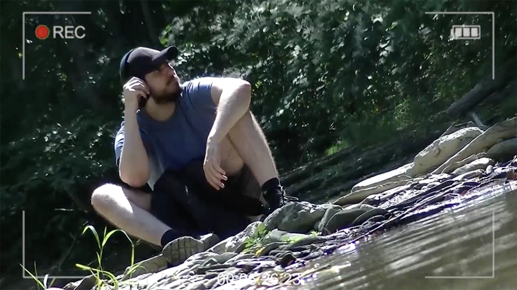 NØKK Movie Thumbnail - Man wearing a hat sitting next to a creek in the forest