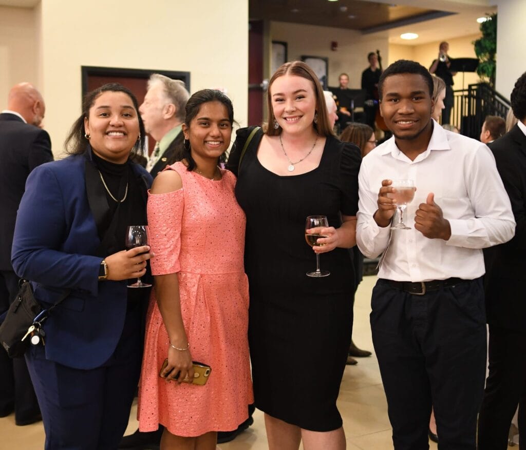 Four students smiling drinking out of wine glasses at a formal party.