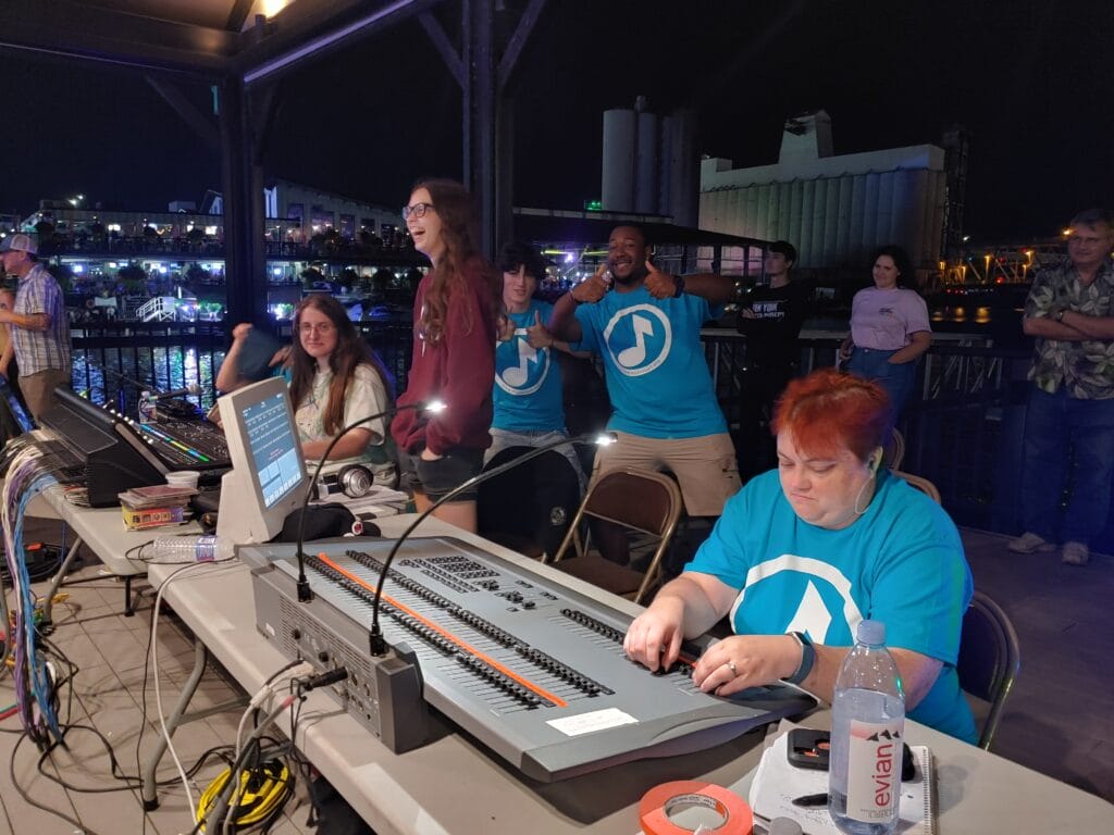 A group of people at an outdoor event at night; two women and a man in blue shirts sit at a control panel, one woman works intently, while others stand behind them, smiling and giving thumbs up. Crowds and lights are in the background.