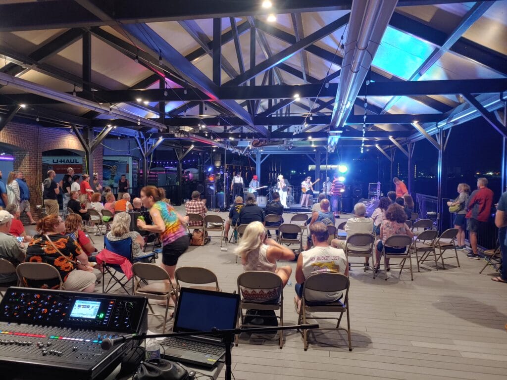 An audience sits on folding chairs under a pavilion at night, watching a live band perform on a lit-up stage. Colorful lights illuminate the scene, and a soundboard is set up in the foreground.