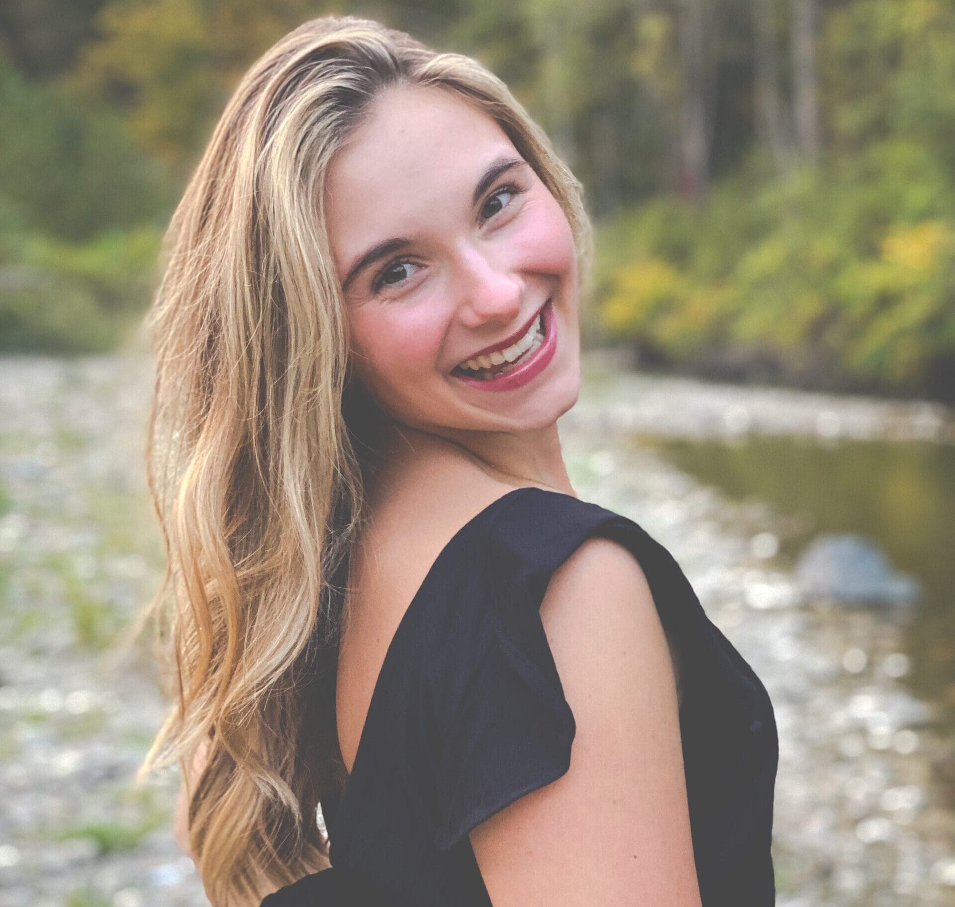 A young woman with long blonde hair, wearing a black dress, smiles while looking over her shoulder. She stands outdoors near a river with trees and rocks in the background.