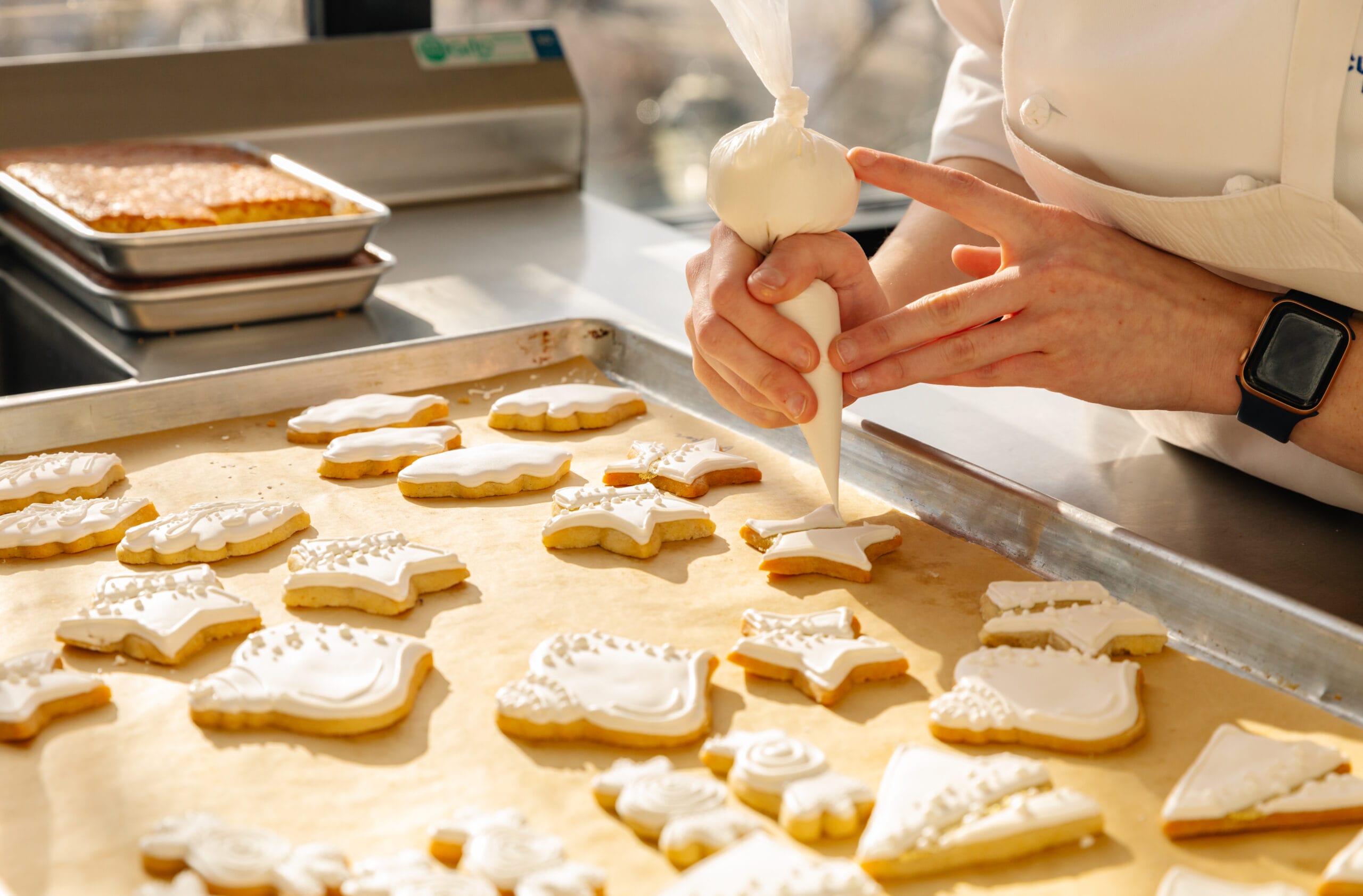 A person wearing a white chef’s coat decorates star-shaped cookies with white icing on a baking tray in a sunlit kitchen. Decorated cookies of various shapes are visible on parchment paper.