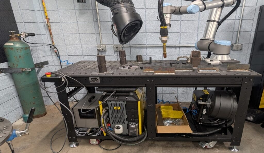 A robotic arm is set up over a metal workbench with various metal pieces and welding equipment. An exhaust hood hangs above, and gas tanks stand nearby. Cables and tools are organized below the work surface.