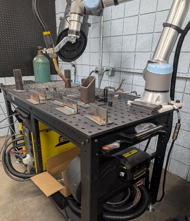 A robotic arm operates over a metal workbench with clamps and metal pieces, in a workshop with a gas cylinder, cables, and welding equipment beneath the table.