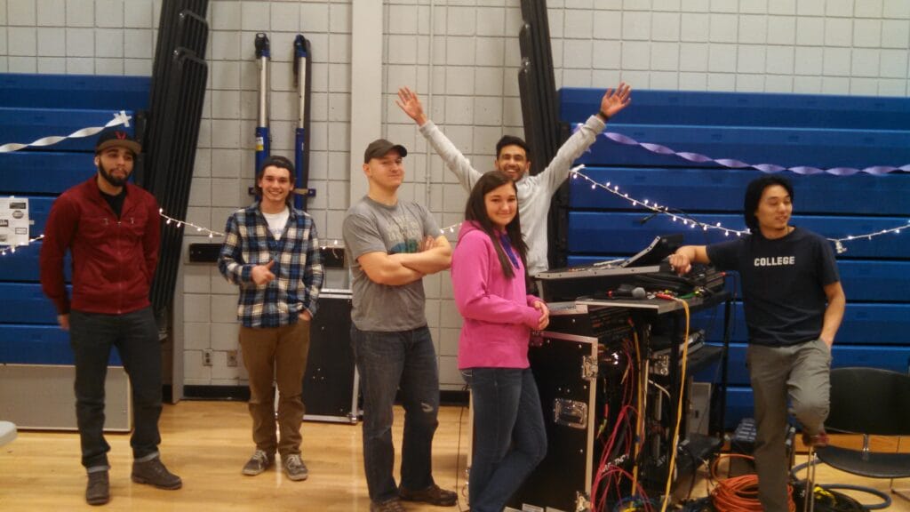 Six people stand in a gymnasium near audio equipment, some smiling and posing. Blue bleachers and white streamers are in the background. One person raises their arms enthusiastically.