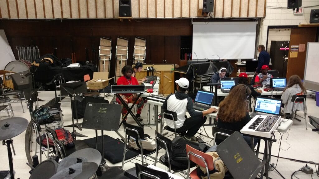 A group of students sits in a music classroom, each working at keyboards and laptops surrounded by music equipment and instruments. An instructor stands near a projector screen at the front of the room.