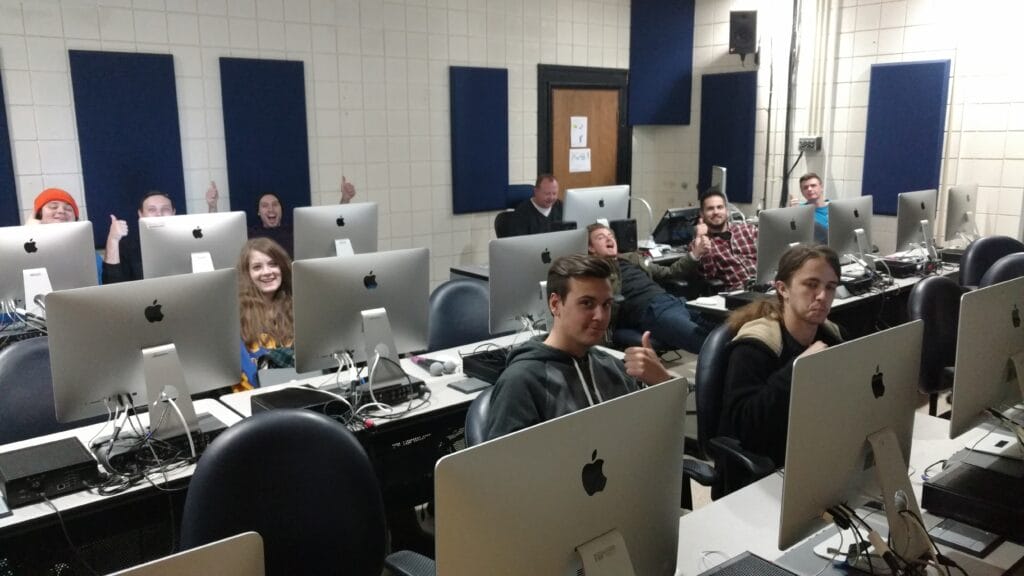 A group of students sit at desks with Apple computers in a classroom. Some smile and give thumbs up, while others look serious or relaxed. The room has white walls, blue panels, and a door at the back.