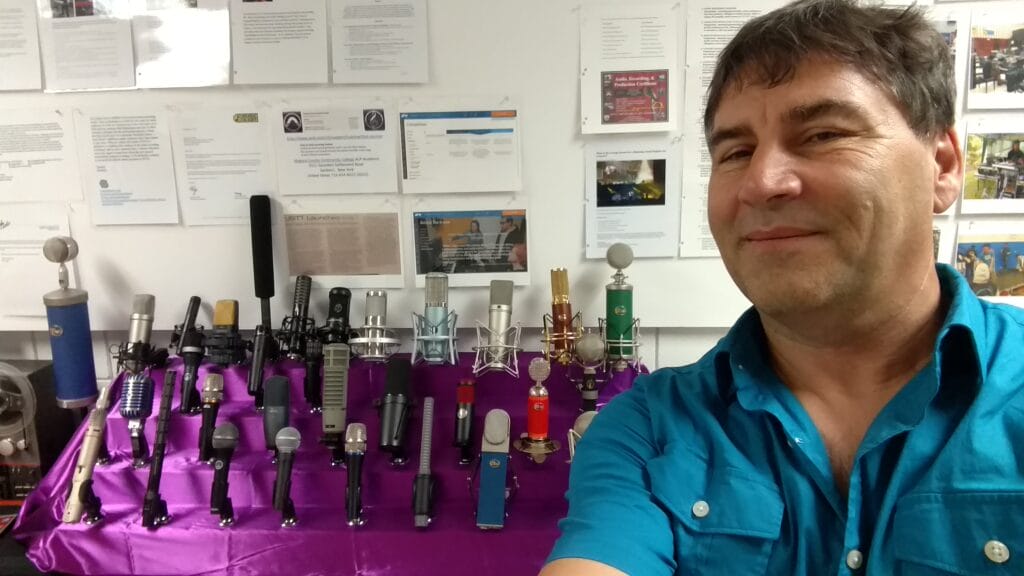 A man in a teal shirt smiles for a selfie in front of a display of various microphones arranged on a purple cloth, with informational posters on the wall behind him.