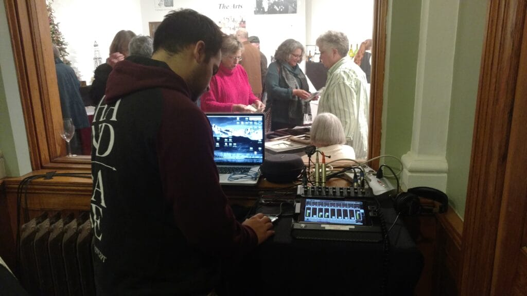 A person operates audio and computer equipment at a table near a window, while a group of people inside a well-lit room interact and browse items on tables.