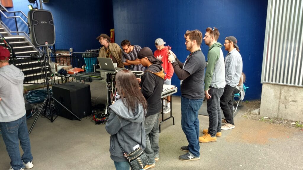 A group of people stands and watches as two men operate DJ equipment on a table under a covered area. Some people are wearing hoodies and hats, and the setting appears to be outdoors near a blue wall.