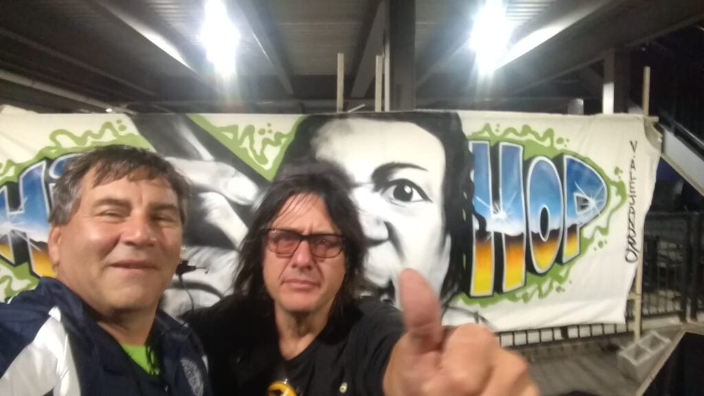 Two men pose for a selfie in front of a colorful mural with the words HIP HOP and a large face. One man gives a thumbs up, and both are smiling under bright lights in an outdoor setting.