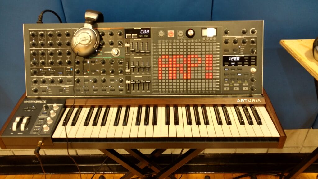 A large Arturia MatrixBrute analog synthesizer with knobs, sliders, buttons, and a digital matrix display sits on a stand. A pair of headphones rests on the top left corner of the synth.