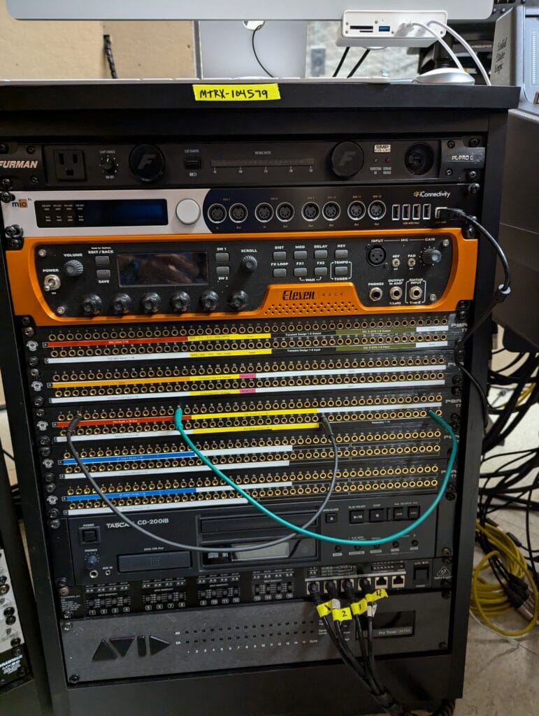 A rack of audio and electronic equipment, including patch panels with many cables plugged in, audio interfaces, and labeled devices. A yellow label with MTRX-194519 is visible at the top of the rack.