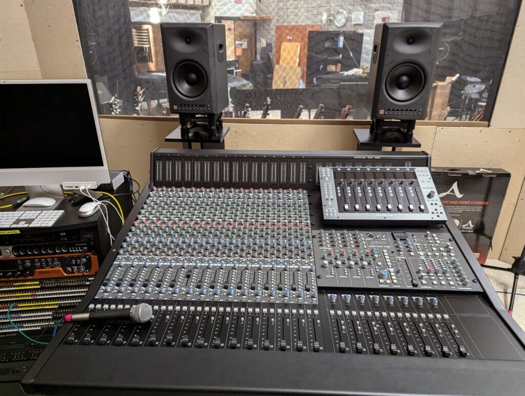 A professional audio mixing console with numerous sliders and knobs, a microphone, speakers, and computer equipment in a recording studio setting, with a window showing another room in the background.