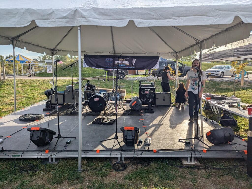 A band setup on an outdoor stage under a white tent, with drums, speakers, and microphones. A person stands at a mic while others set up equipment. Grass, cars, and a blue sky are visible in the background.