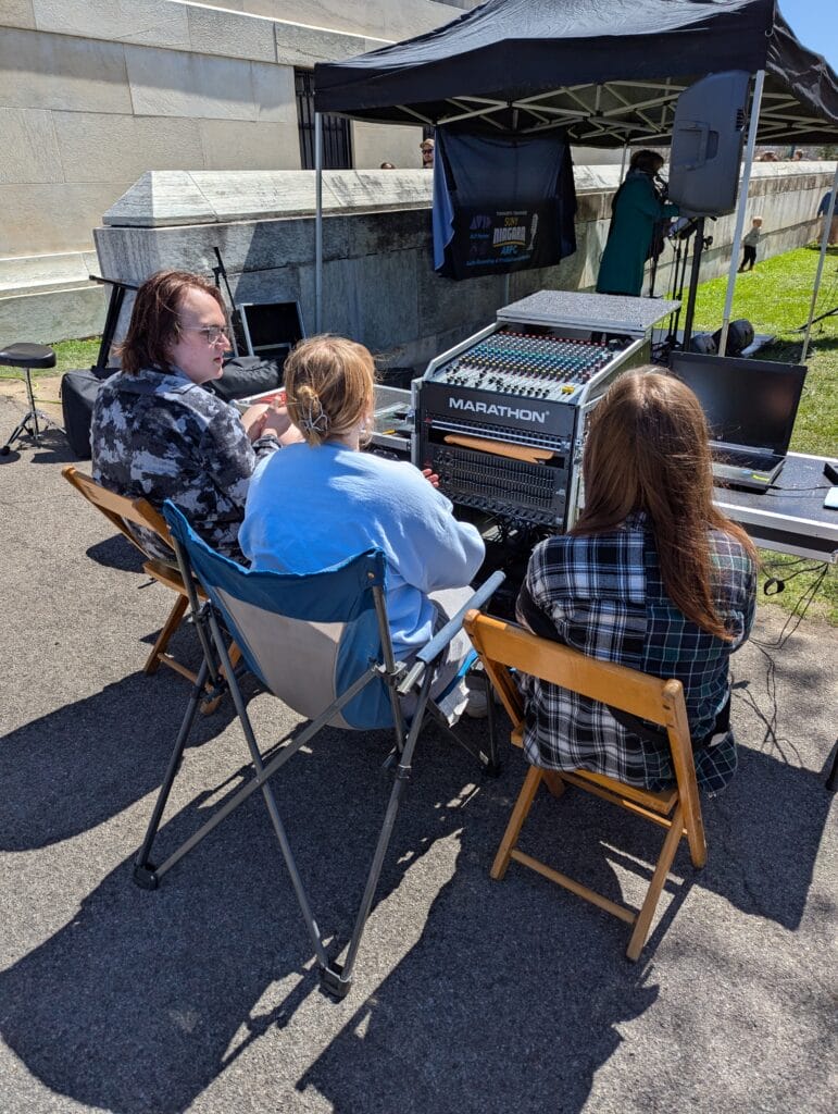 Three people sit on folding chairs outside at a sound mixing station with audio equipment and laptops, under a tent on a sunny day. A stone wall and green grass are in the background.