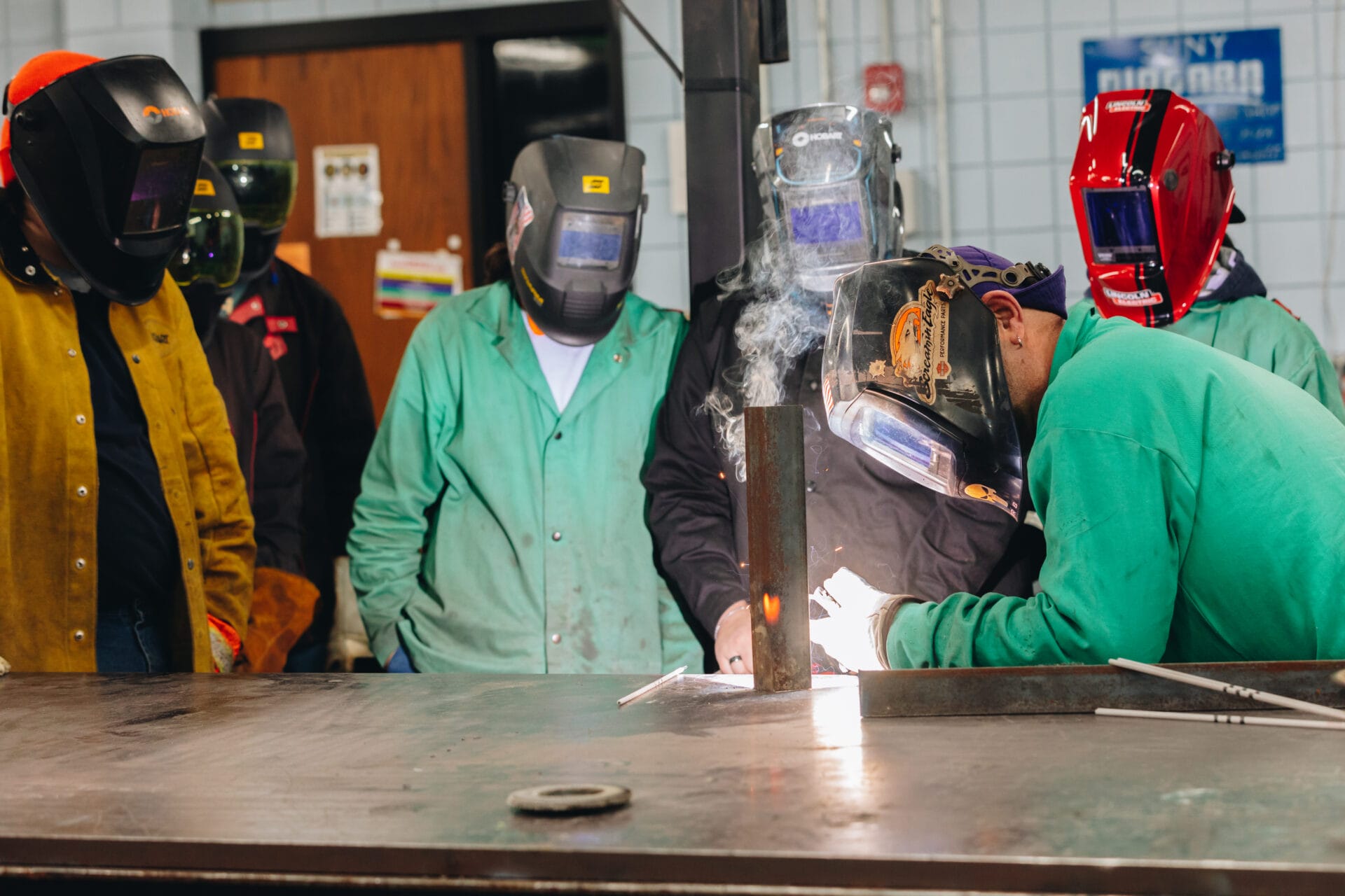 A group of people wearing protective welding gear and helmets watch closely as one individual welds metal on a worktable—showcasing Workforce Development at SUNY Niagara: Empowering the Community Through Skilled Training and Certification Programs.