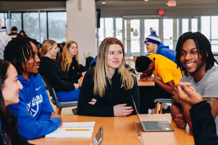 A group of college students sit around a table in a common area, talking and smiling, with laptops and notebooks in front of them. More students are visible in the background, engaged in conversation and studying.
