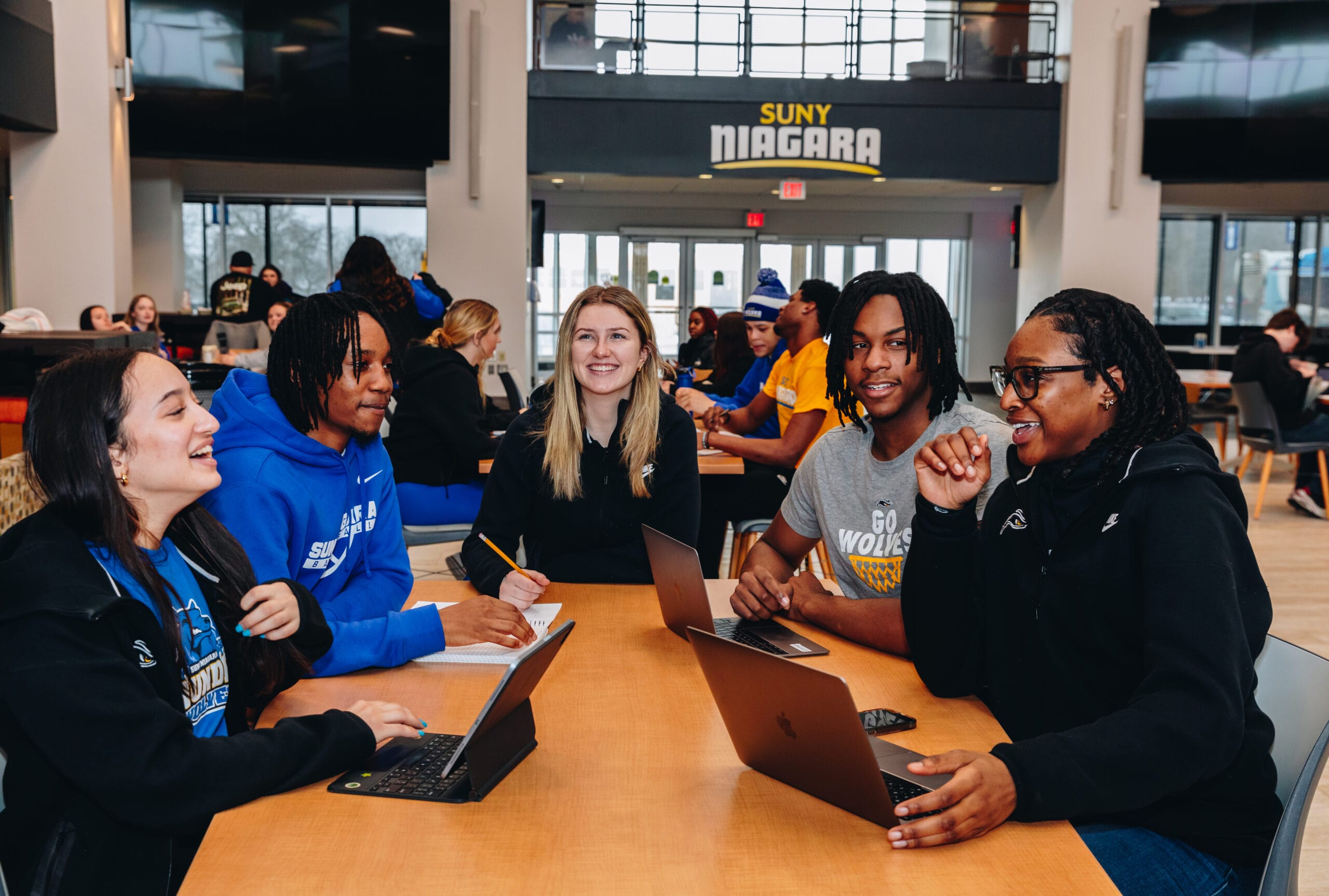 Five students sit around a table in a SUNY Niagara campus common area, smiling and talking while working on laptops and notebooks; other students socialize in the background. Discover vibrant campus life—apply to SUNY Niagara today!.