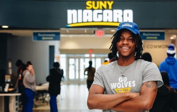 A smiling person wearing a blue Niagara cap and a gray Go Wolves t-shirt stands with arms crossed inside a SUNY Niagara building, with people and signs visible in the background.
