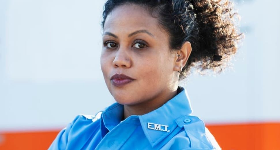 A woman in a blue EMT uniform stands with her arms crossed, looking confidently at the camera. An ambulance is visible in the background.