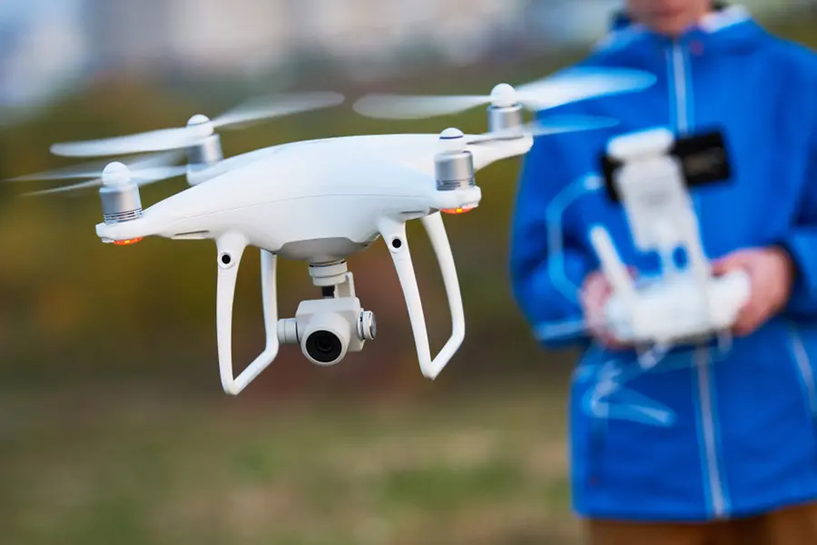 A white quadcopter drone with a camera hovers in the air, while a person in a blue jacket operates it using a remote controller in the background.