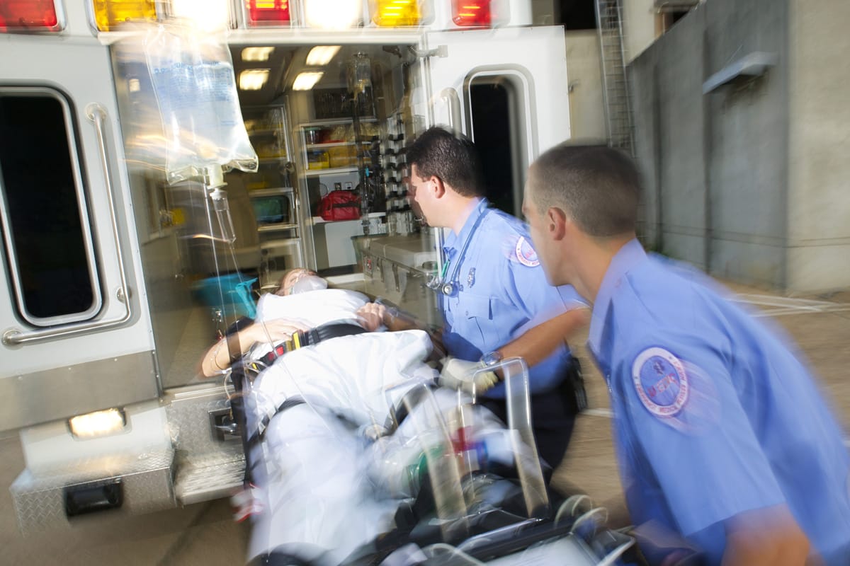 Two paramedics in blue uniforms rush a patient on a stretcher into an ambulance, with medical equipment visible inside the vehicle and a sense of urgency in their movements.