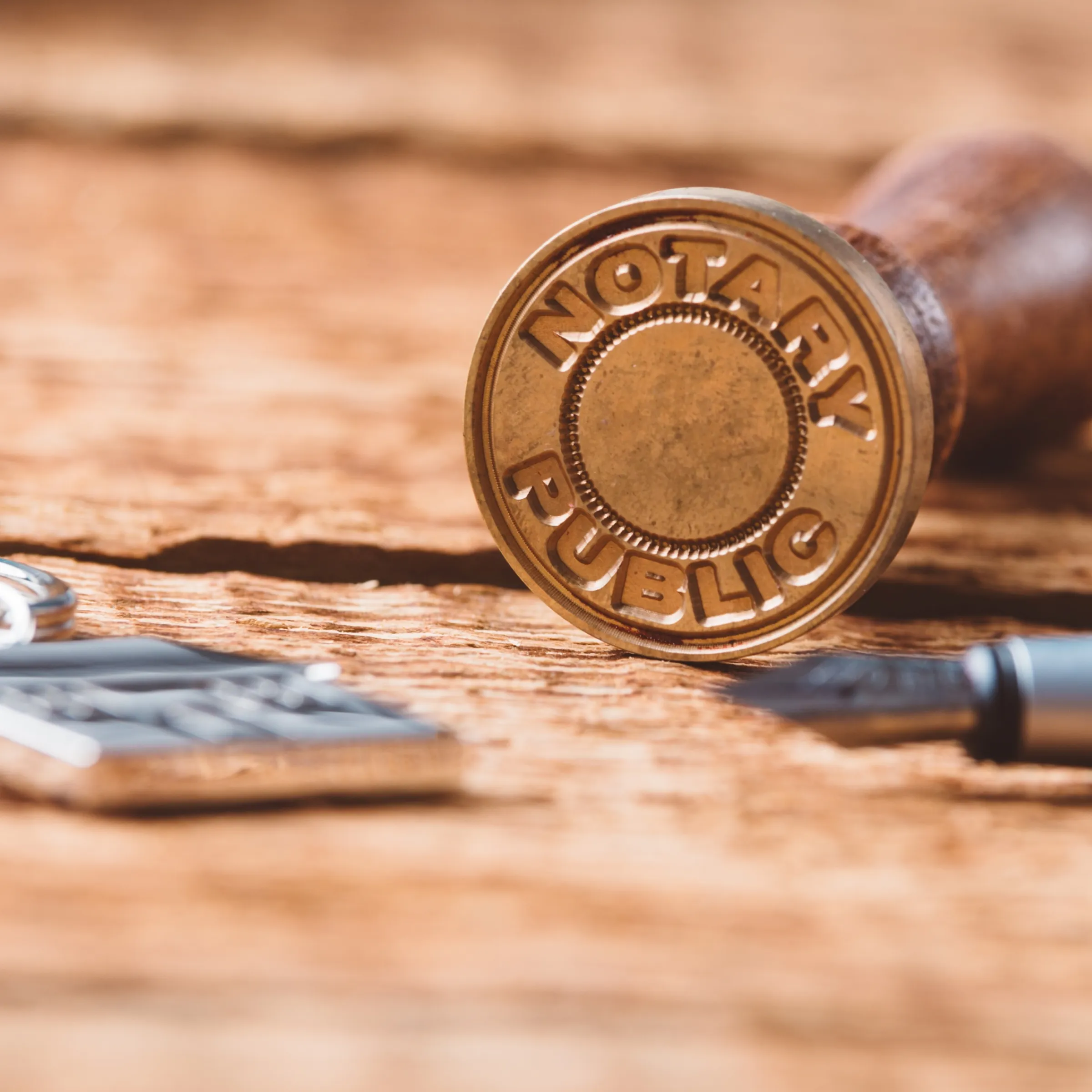 A notary public stamp lying on a wooden surface next to a fountain pen and a keychain. The stamp’s seal is visible, displaying the words “Notary Public” in raised letters.