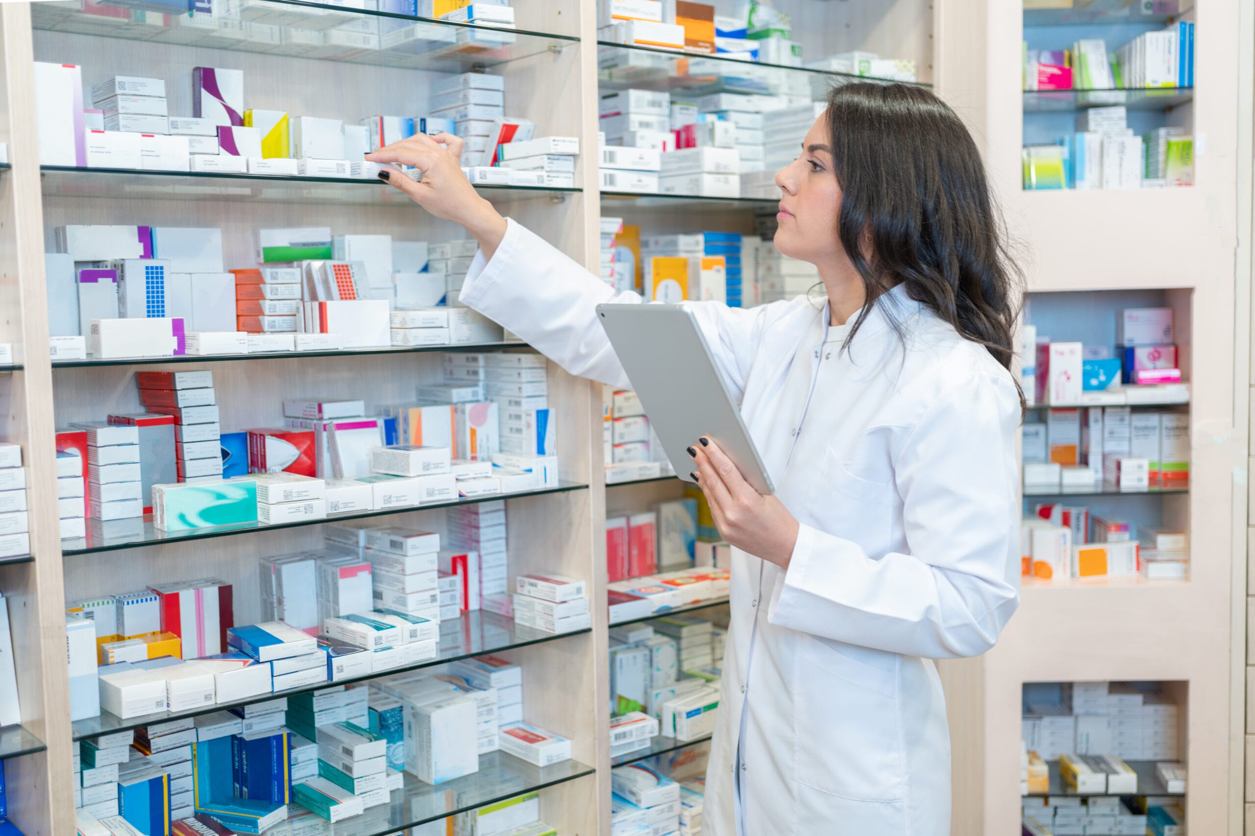 A pharmacist in a white coat holds a digital tablet while reaching for a box of medication on a shelf filled with various medicine boxes in a pharmacy.