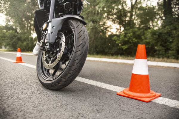 Close-up of a motorcycle front wheel on an asphalt road, maneuvering between orange and white traffic cones, with green trees visible in the background.