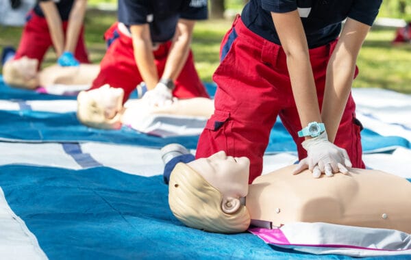 People wearing red pants and gloves practice CPR on training mannequins outdoors, pressing on the mannequins’ chests while kneeling on blue mats.
