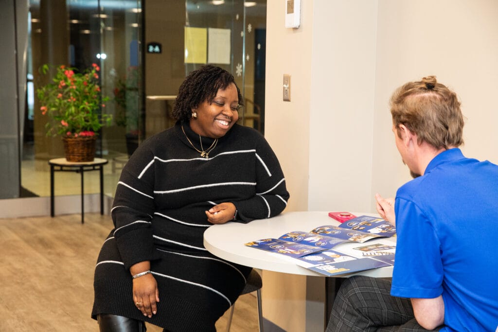 A woman in a black striped dress smiles while seated at a round table with a man in a blue shirt, who is holding brochures. Indoors at night, a potted plant is visible in the background.
