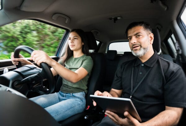 A young woman drives a car while an instructor sits beside her holding a tablet, appearing to give directions or feedback during a driving lesson. Trees are visible outside the window.