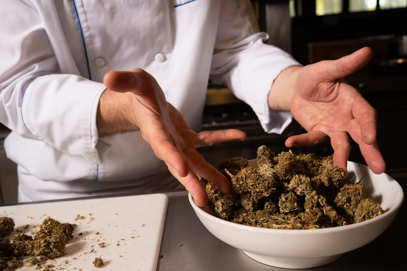 A chef in a white coat gestures over a large white bowl filled with cannabis buds on a kitchen counter, with more buds on a cutting board nearby.