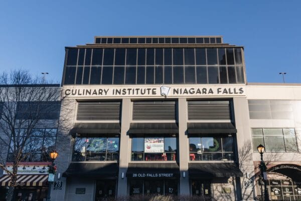 Photo of the front of Niagara Falls Culinary Institute which has black windows and a chefs hat logo angled up Infront of a blue, bright, sunny sky.