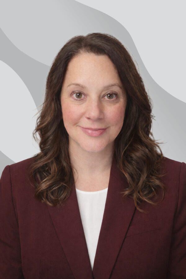 A woman with long brown hair, wearing a maroon blazer and white top, smiles at the camera in front of a gray abstract background.