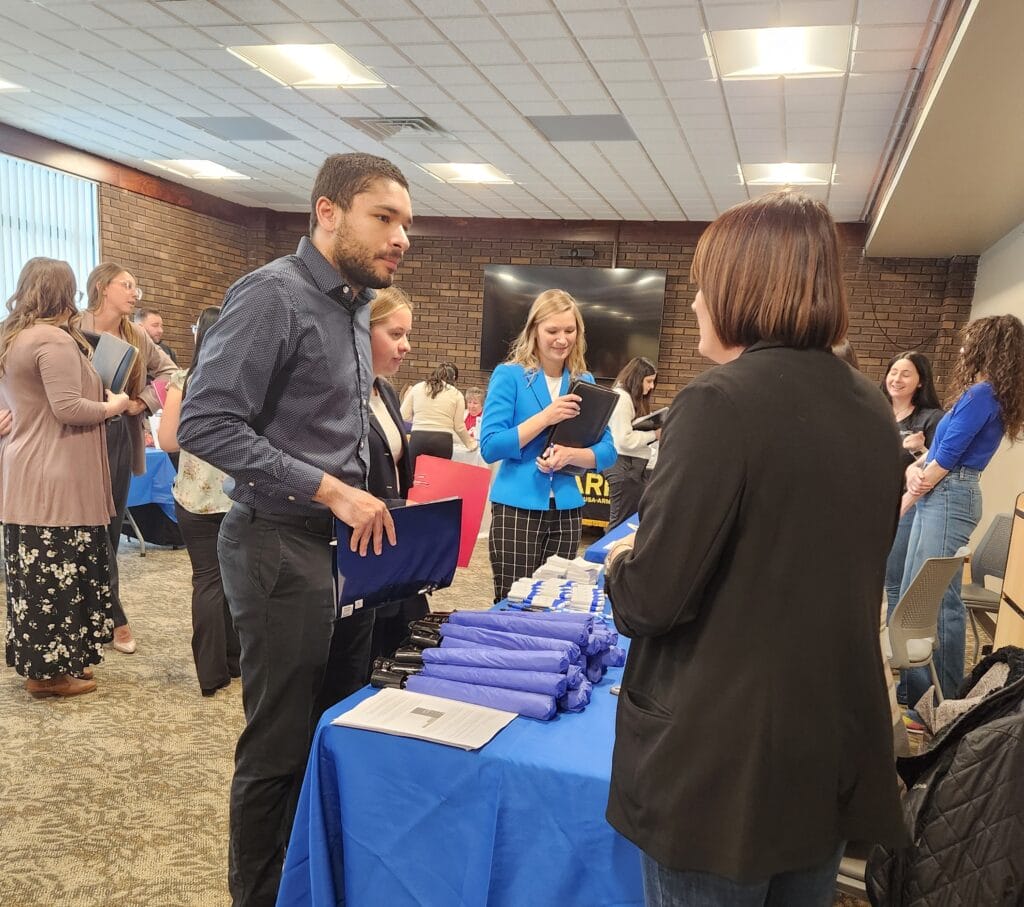 A group of people conversing indoors at what appears to be a fair or presentation event. In the foreground, a young man holding a portfolio or folder stands facing a woman whose back is to the camera.