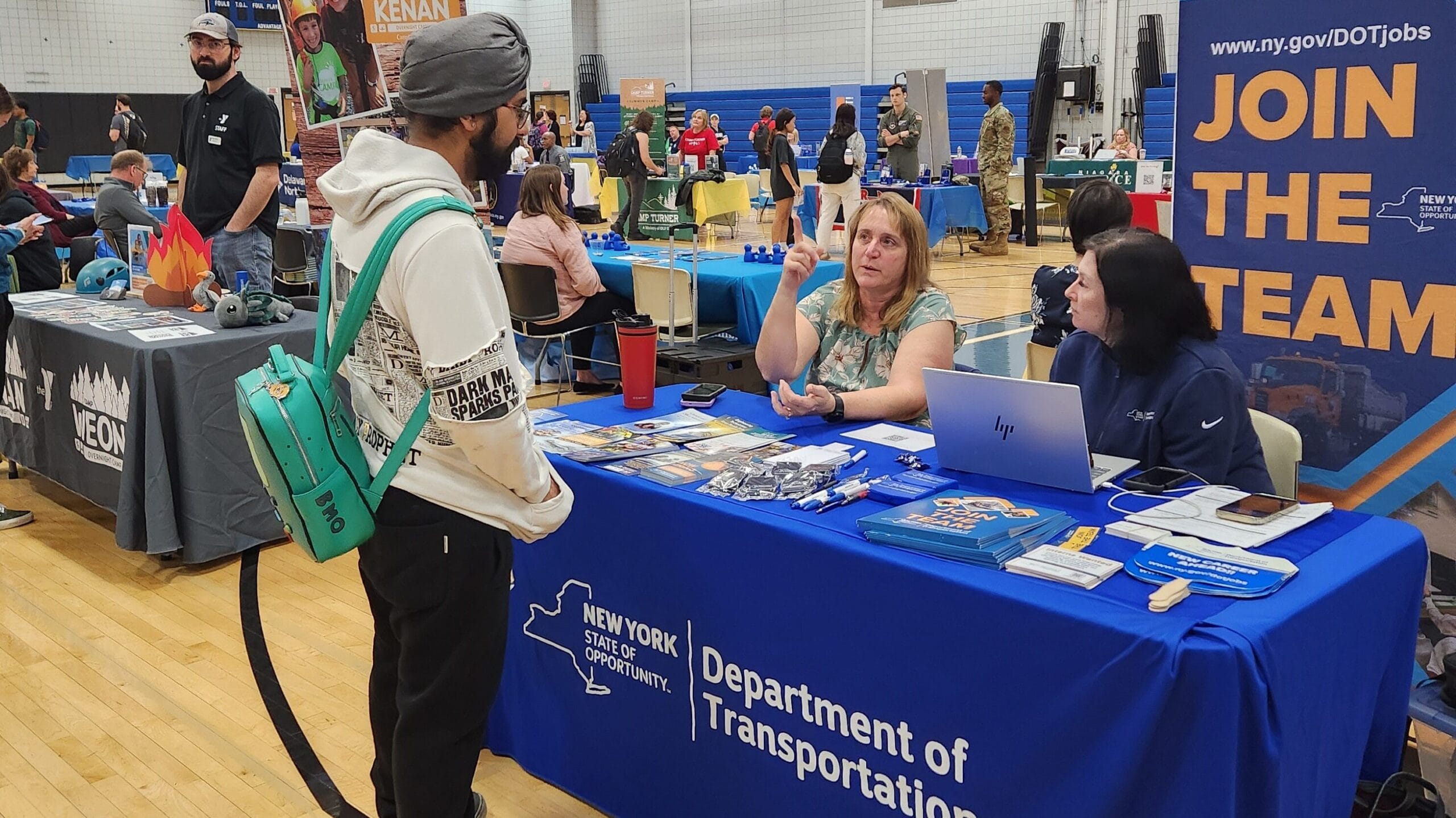 A well-lit gymnasium or indoor event space set up with tables for an informational fair. In the foreground, a young man in a turban and white hoodie, carrying a teal backpack with 'BMO' on it, stands at a table draped in a blue cloth labeled ‘New York State of Opportunity—Department of Transportation.’ On the table are brochures, pens, and a laptop. Behind the table, two representatives—one gesturing with her hand and the other seated—appear to be explaining something. To the right, a tall banner reads ‘Environmental · Engineering · Analyst · Legal · Admin · Technician’ and ‘Join the Team’ with a URL for DOT job opportunities. In the background, more tables and attendees are visible across the gymnasium floor.